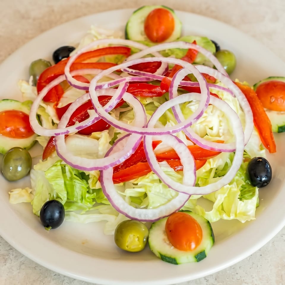 A fresh garden salad with lettuce, red onions, bell peppers, cucumbers, and olives on a white plate.