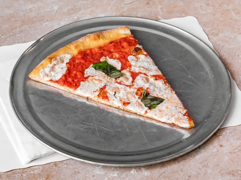 A single slice of pizza with red sauce, white cheese, and basil on a metal tray.